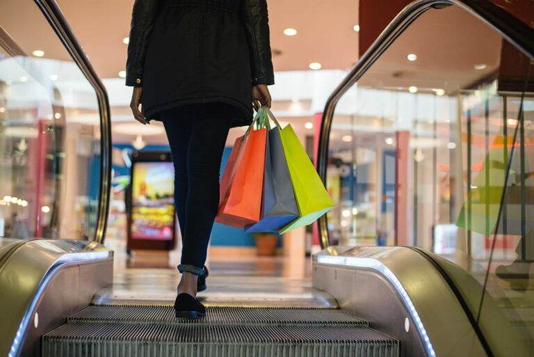 A woman holding colourful shopping bags walks off an escalator