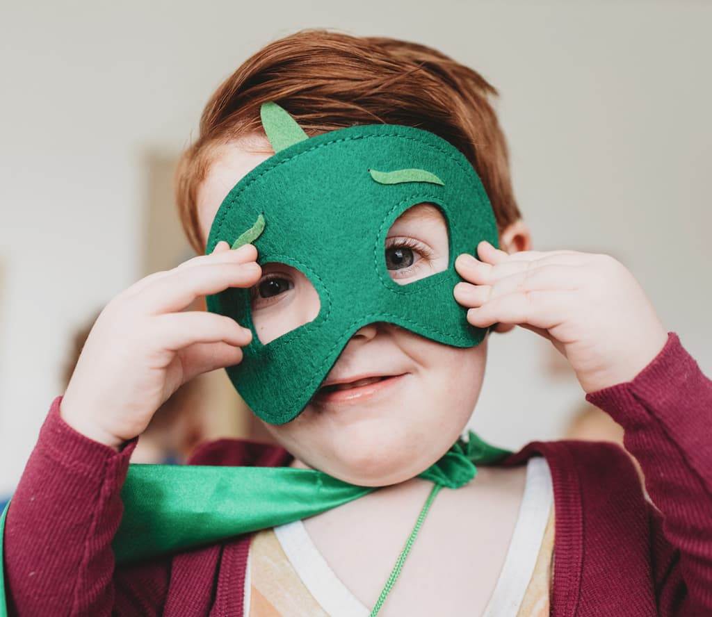A child holding a felt mask to their face and wearing a cape