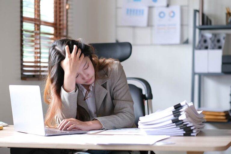 A businesswoman sits at a desk with piles of paperwork holding her head in her hand